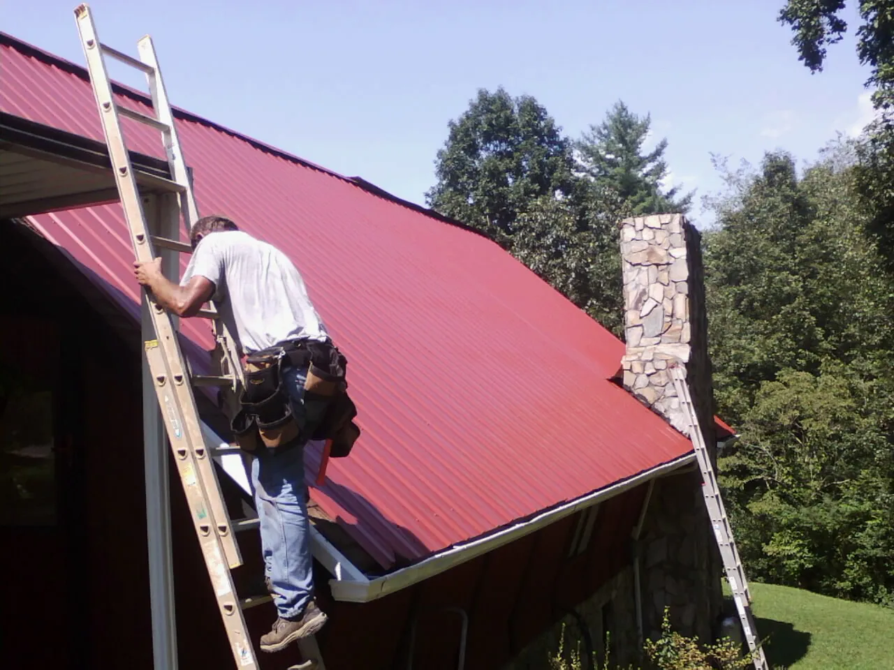 Roofer on a ladder beside a red metal roof and stone chimney — Soffit & Fascia Repair by Roofing Brothers in Stanfield